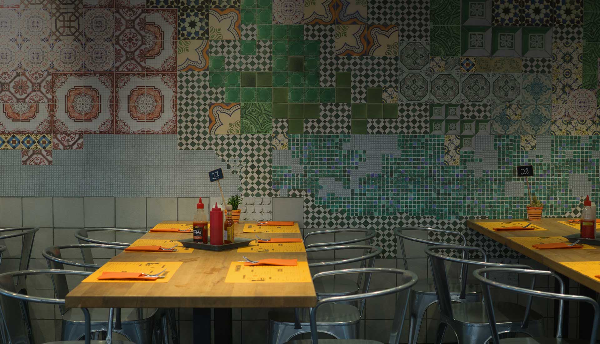 Wnętrze restauracji z drewnianymi stołami, metalowymi krzesłami i kolorową ścianą wyłożoną kafelkami.